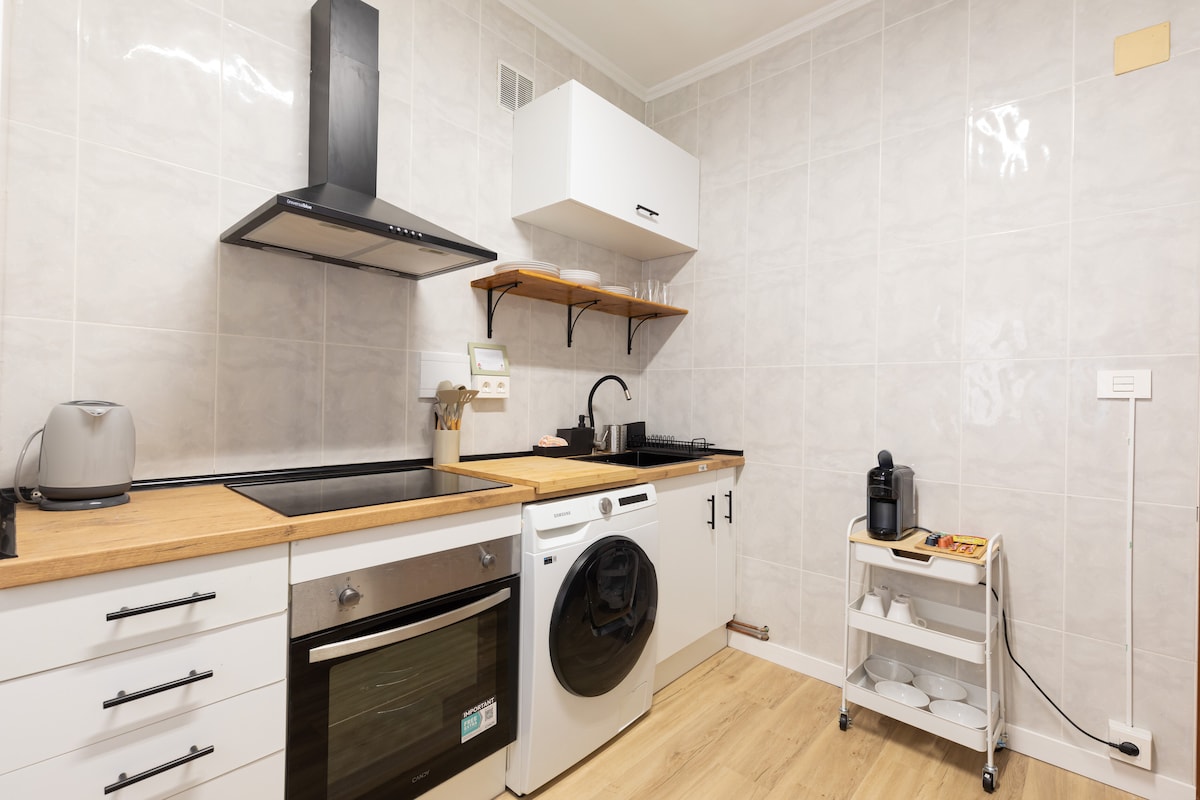 A modern kitchen is equipped with essential appliances, including an electric kettle, stovetop, and dishwasher. Wooden countertops provide a warm contrast to the sleek white cabinetry. A small cart holds kitchen items, enhancing the practical layout of the space.