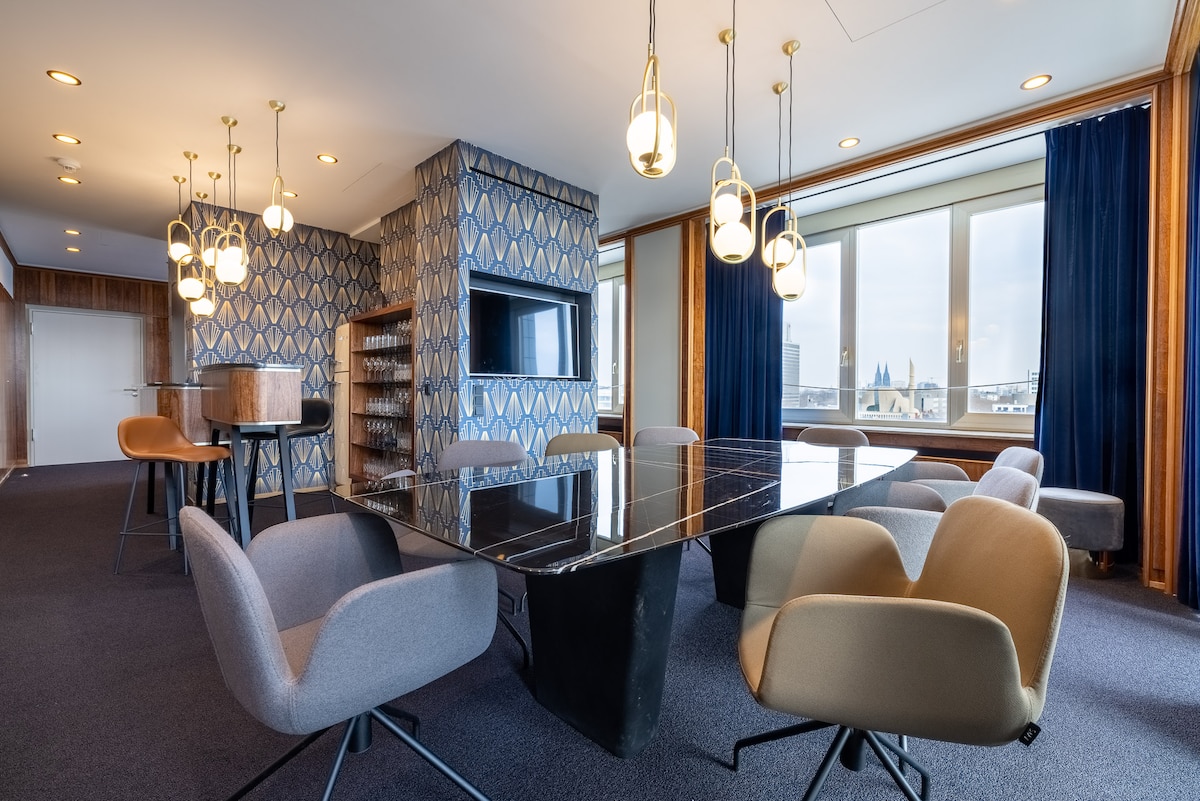 A spacious communal area features a large dining table surrounded by modern chairs. Elegant pendant lights hang from the ceiling, illuminating the space. Floor-to-ceiling windows provide scenic views, while blue drapes complement the contemporary decor, creating a comfortable and inviting setting.