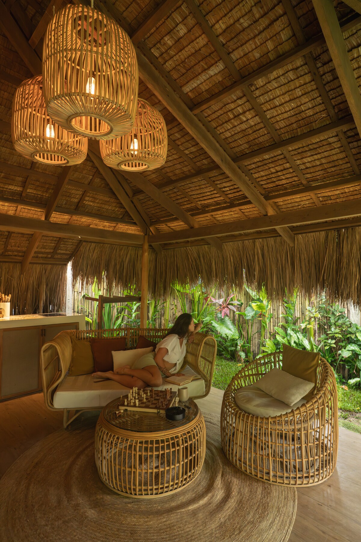 A natural, bamboo-style seating area is framed by lush tropical foliage. Two woven chairs are complemented by a round table, adorned with a tray and glassware. A statement chandelier hangs from the thatched ceiling, casting a warm glow over the inviting space.
