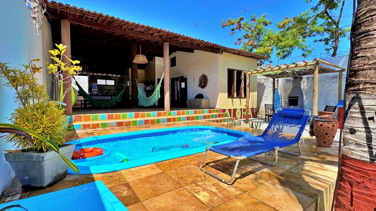 A private outdoor area features a swimming pool bordered by a tiled deck, with lounge chairs positioned for relaxation. Above, a shaded patio with a traditional roof complements the space, while vibrant greenery enhances the inviting atmosphere.