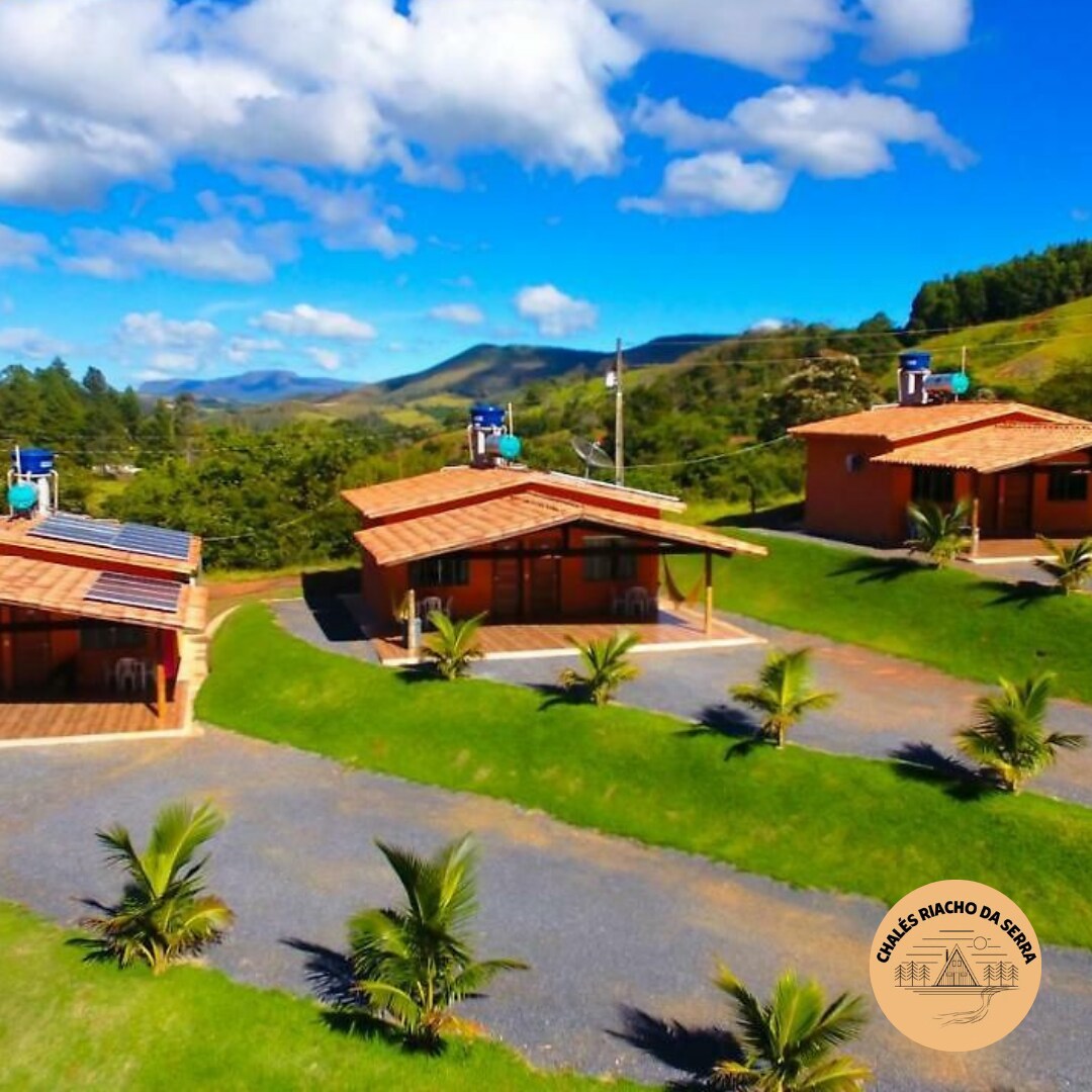 Chalets are nestled in a lush green landscape, surrounded by rolling hills and clear blue skies. Each chalet features a wooden structure with a tiled roof, set against a backdrop of palm trees and well-maintained grass paths.