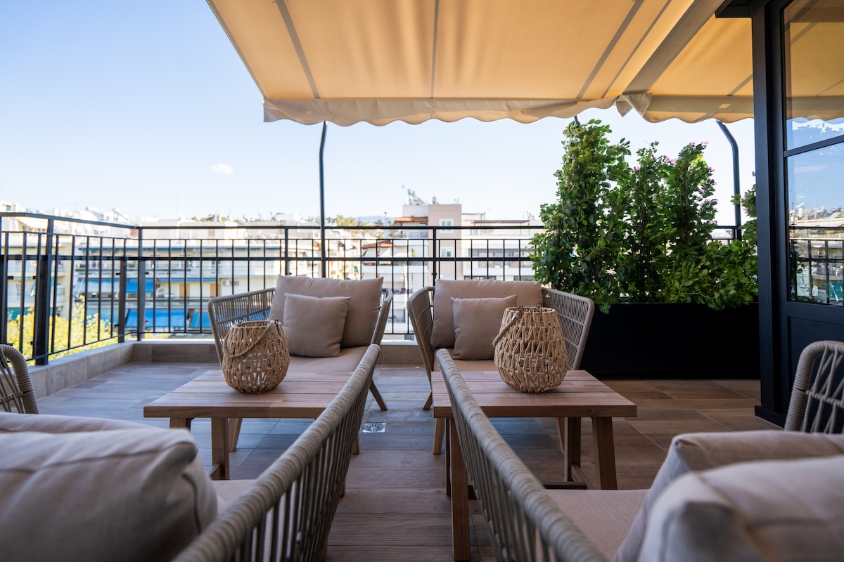 A spacious terrace features elegant patio furniture arranged around wooden tables. The area is shaded by a retractable awning, with potted greenery adding a touch of nature. A railing provides safety while overlooking the surrounding views.