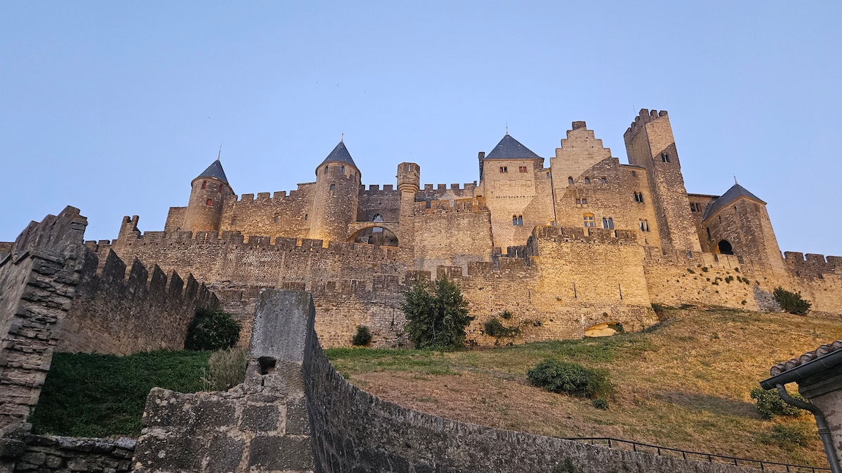 The medieval fortifications of Carcassonne rise majestically, showcasing tall stone walls and towers under a softly lit evening sky. The castle's structured silhouette is complemented by surrounding greenery, creating a striking contrast. The image captures the historical architecture and grandeur of this iconic landmark.