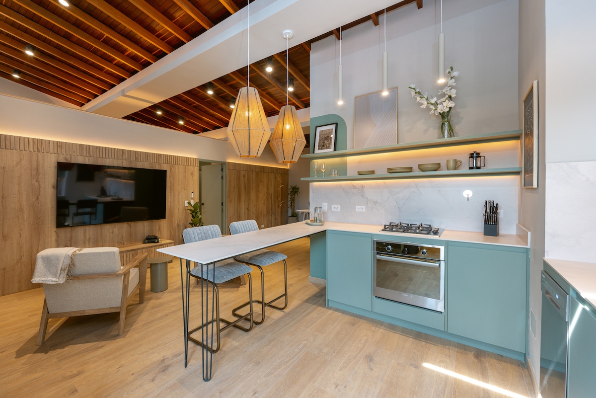 A modern kitchen area features light wood accents and teal cabinetry. Two high stools are positioned at a countertop, complemented by pendant lights overhead. An equipped stove and wall-mounted cabinetry provide functional space. The room showcases a cozy seating arrangement and a large television mounted on a wooden wall.