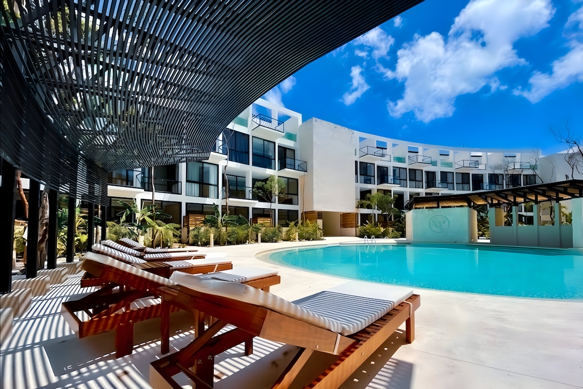 A serene pool area features a turquoise water surface, surrounded by relaxing lounge chairs. The modern building showcases multiple balconies and lush greenery, creating a calm retreat. Soft sunlight filters through an overhead slatted shade, enhancing the tranquil ambiance.