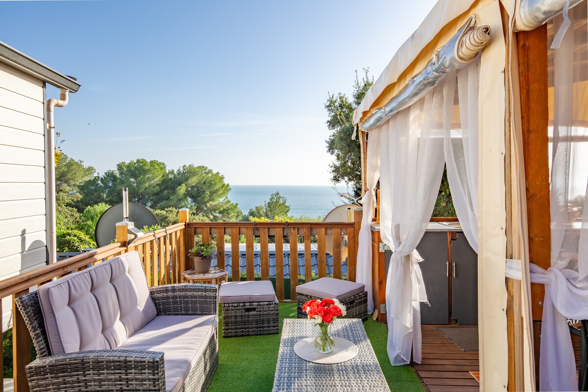 A terrace with a view of the sea features a cozy seating area, including a framed shelter and lush green flooring. Soft, natural light illuminates the space, enhancing a sense of relaxation. A small table holds a vase with vibrant flowers, adding a touch of color.