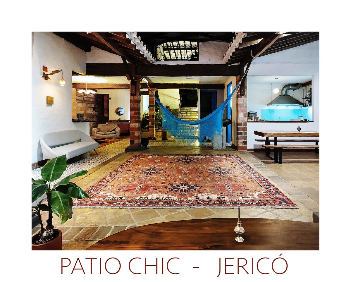 An inviting interior space showcases a large area rug in warm tones, complemented by a light-colored sofa and a blue hammock. Exposed wooden beams and natural stone flooring contribute to a relaxed ambiance, while an indoor water feature is visible in the background.