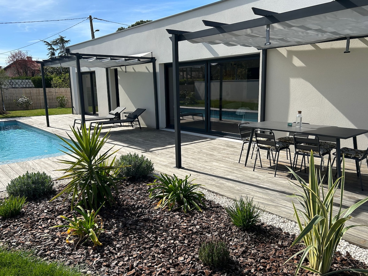 A sunlit terrace features a swimming pool and a dining area with a table and chairs. Two lounge chairs are positioned beside the pool. Decorative plants are arranged around the terrace, enhancing the outdoor space's natural appeal.