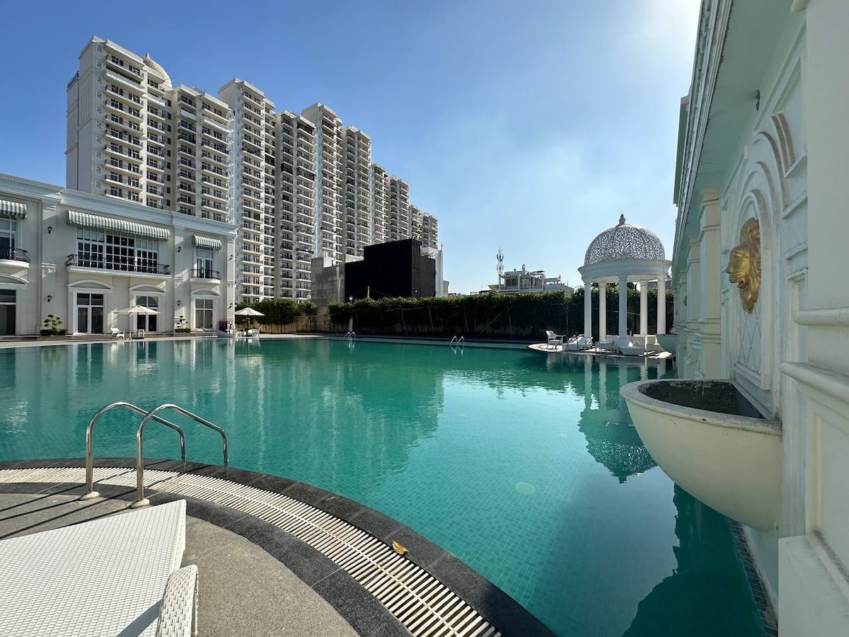 A serene swimming pool with crystal-clear turquoise water is featured, surrounded by modern buildings. A white ornate gazebo stands at one edge, suggesting a tranquil area for relaxation. Comfortable lounge chairs are positioned near the pool, creating an inviting space for guests.