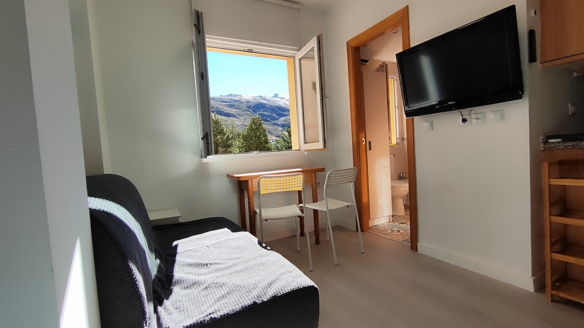 A cozy seating area features a dark sofa positioned beside a window, allowing views of the surrounding mountains. A small wooden table is paired with two white chairs. A mounted television is visible on the wall, adding to the functionality of the space.