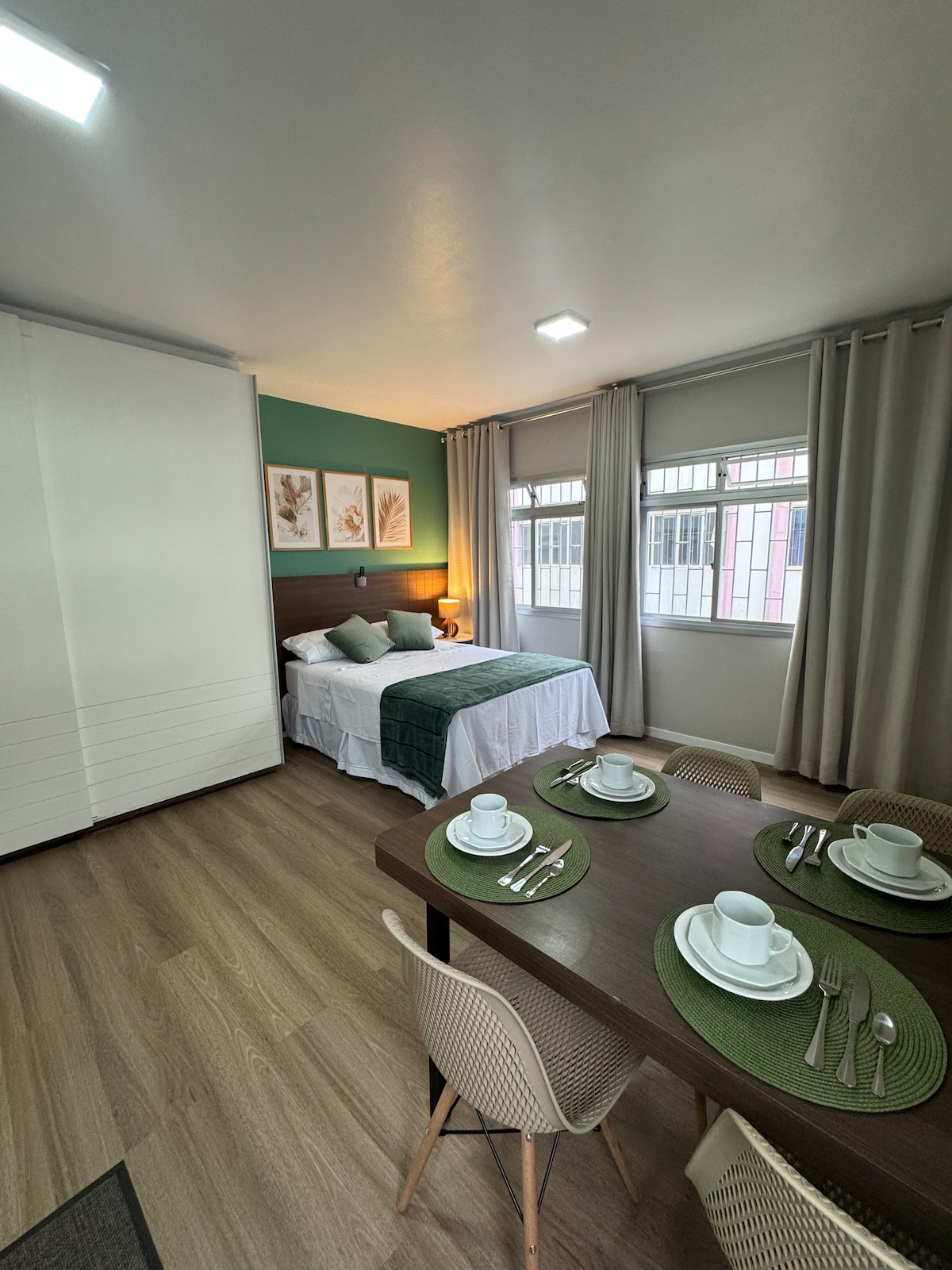 A well-lit studio features a spacious bed with a green throw and decorative wall art. A dining table is set for four, showcasing white dishware and utensils. Large windows dressed with light curtains allow natural light to brighten the inviting space.