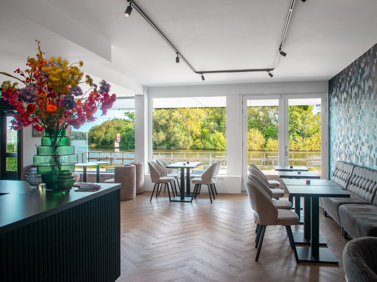 The spacious dining area is adorned with elegant furnishings, featuring light-colored chairs and small tables. A large floral arrangement in a green vase adds a pop of color. Large windows offer natural light and views of the surrounding greenery by the lake.