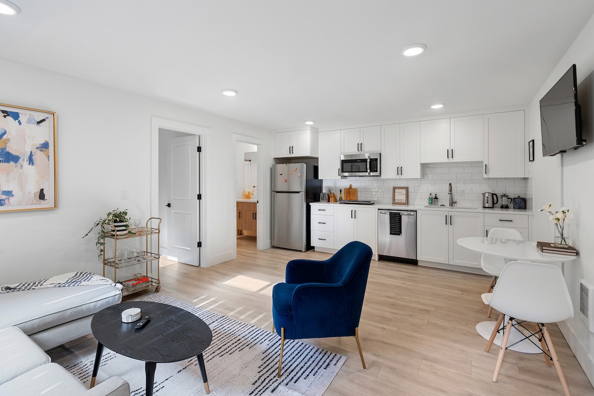A bright living area features modern furnishings, including a blue accent chair and a circular black coffee table. The open kitchen is equipped with white cabinets, stainless steel appliances, and a dining table. Natural light floods the space through the windows, enhancing the welcoming environment.