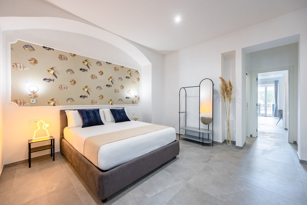 A spacious bedroom features a cozy bed with a softly upholstered headboard, complemented by playful fish-patterned wallpaper. A stylish lamp is positioned next to a minimalist nightstand. Smooth gray tiles extend across the floor, enhancing the room's modern aesthetic and inviting atmosphere.