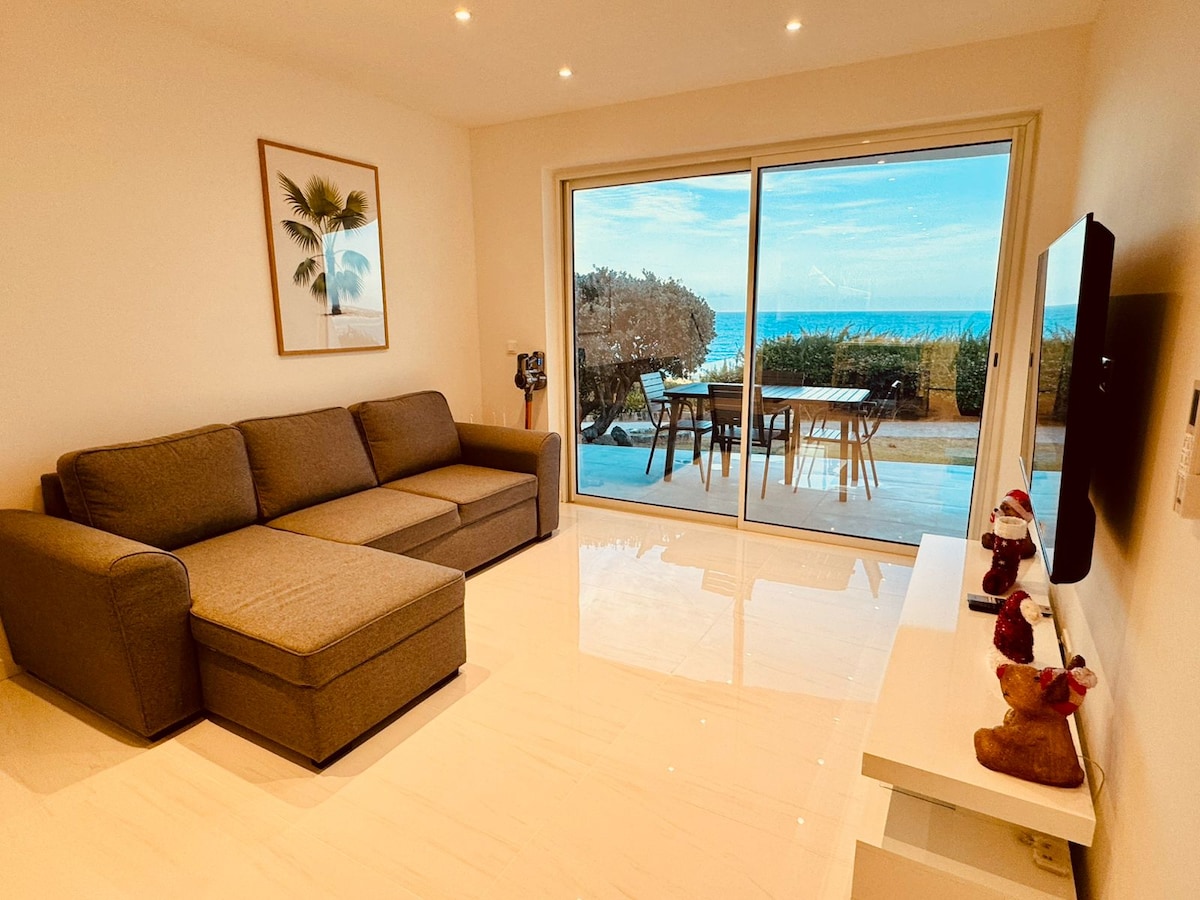 A spacious living area features a comfortable sectional sofa positioned near large sliding glass doors, offering a view of the outdoor patio and sea. A flat-screen television is mounted on the wall, and natural light illuminates the glossy tile floor.