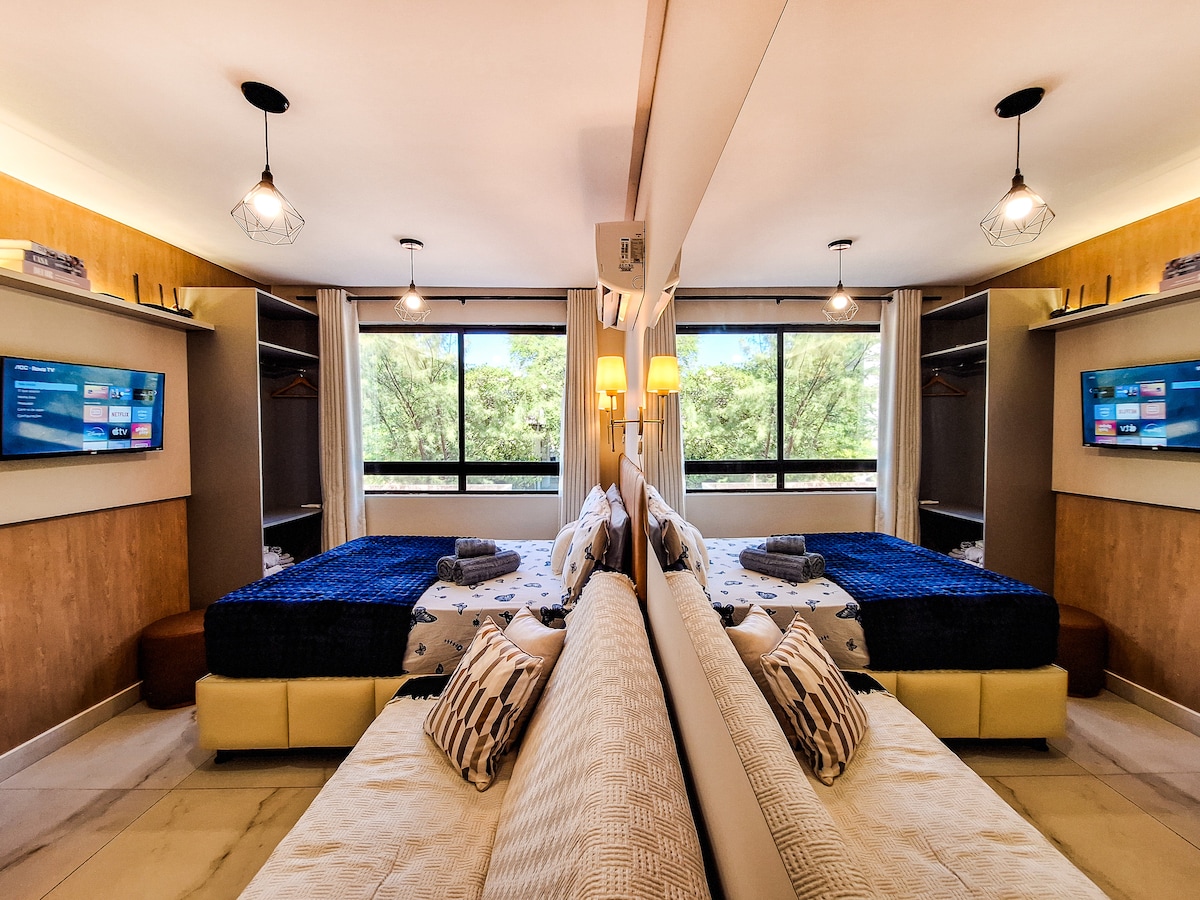 A well-appointed sleeping area features two spacious beds, each adorned with neatly arranged linens. Natural light streams through large windows, providing a view of greenery outside. Modern decorative elements and accent pillows enhance the inviting atmosphere, while built-in shelves offer convenient storage solutions.