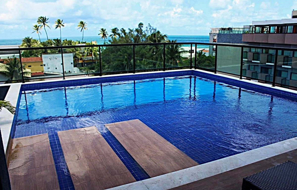 A rooftop pool offers panoramic views of the ocean and surrounding palm trees. The tranquil water reflects the sky, while wooden decking provides a space for relaxation. The area is enclosed by glass railing, enhancing the openness of the setting.