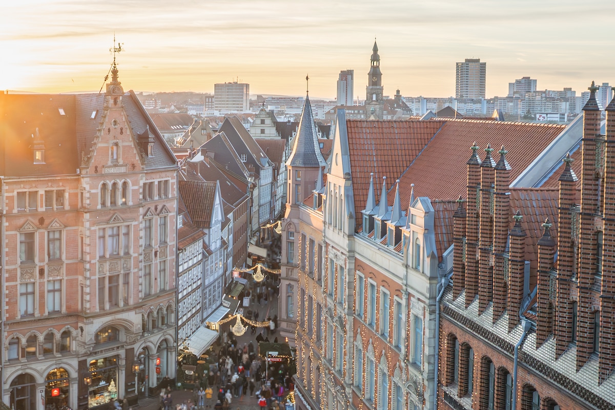 A view of Hannover's rooftops is captured during sunset, with sunlit historical buildings showcasing intricate architectural details. The skyline in the background features modern structures alongside traditional styles, creating a blend of old and new in the cityscape.