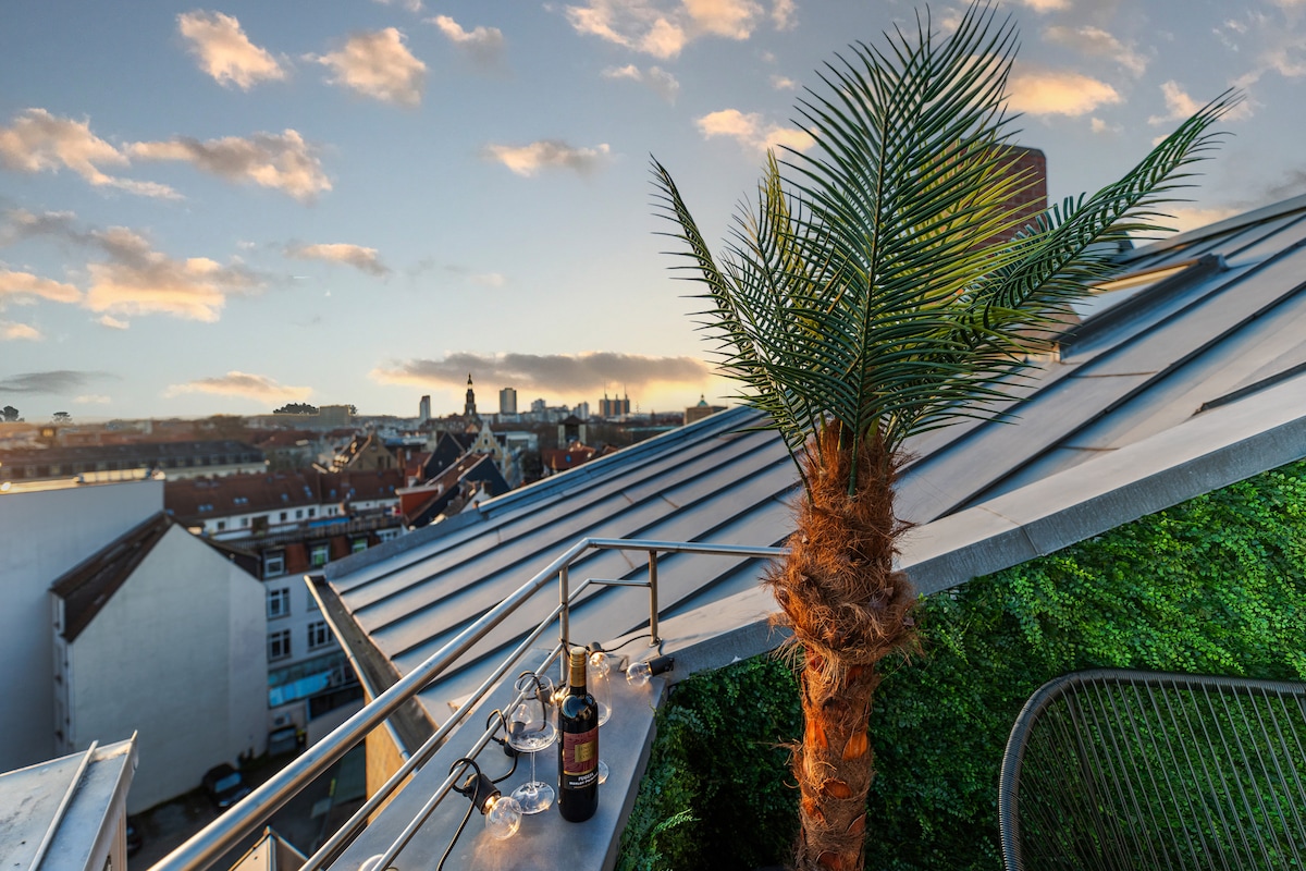 A rooftop terrace is depicted, featuring a palm tree and a bottle of wine with glasses arranged on a table. The skyline of Hannover is visible in the background, framed by a vibrant sunset sky filled with scattered clouds.
