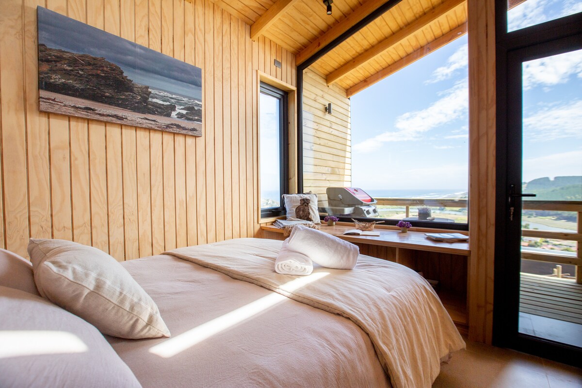A bright bedroom features warm wooden walls and large windows that offer views of the outdoors. A spacious bed with neutral bedding is complemented by rolled towels. A small table is positioned beside the bed, showcasing a magazine and additional items.