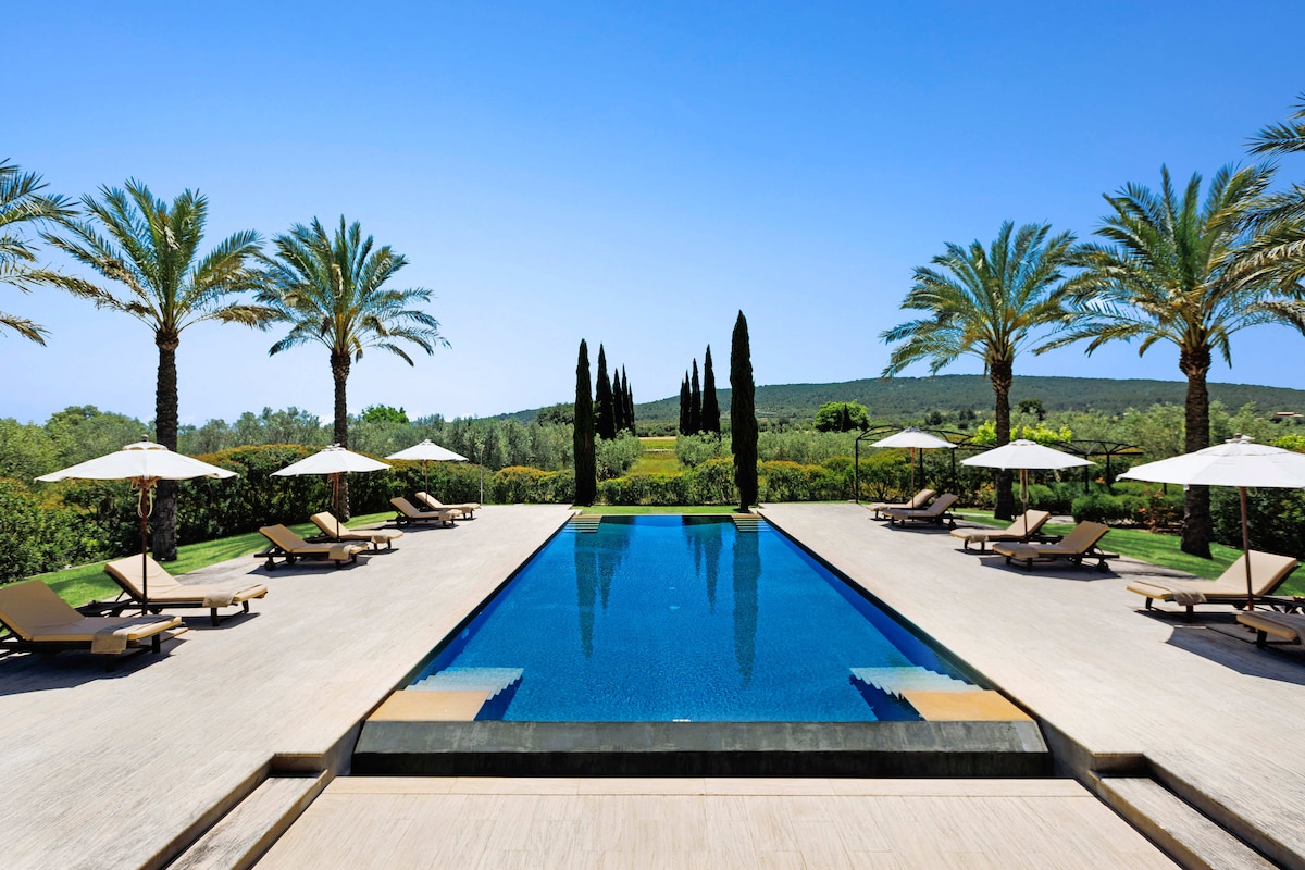 A spacious pool area is framed by palm trees, featuring multiple lounge chairs arranged neatly under large umbrellas. The clear blue water of the pool reflects the bright sky, while manicured hedges and cypress trees enhance the scenic view in the background.