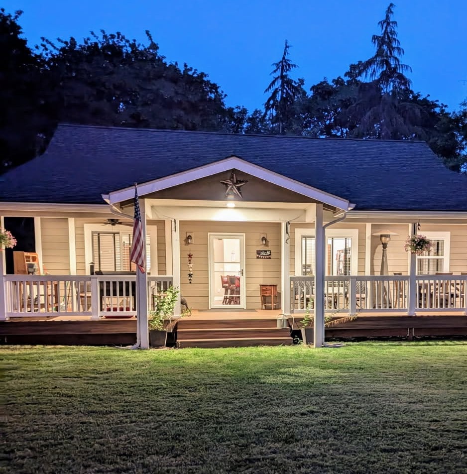 The front exterior of the house is illuminated in the evening light, featuring a welcoming porch with decorative elements. An American flag hangs to the right of the entrance, while potted plants add greenery. A spacious lawn provides an attractive setting.