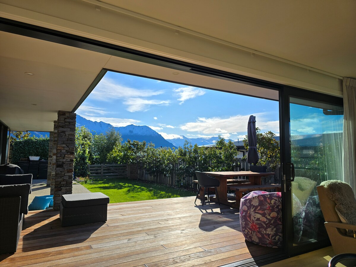 A spacious outdoor area is visible, featuring a wooden deck with a dining table and lounge seating. Large glass doors open to the breathtaking view of distant mountains and lush greenery, creating a seamless transition between indoor and outdoor spaces.