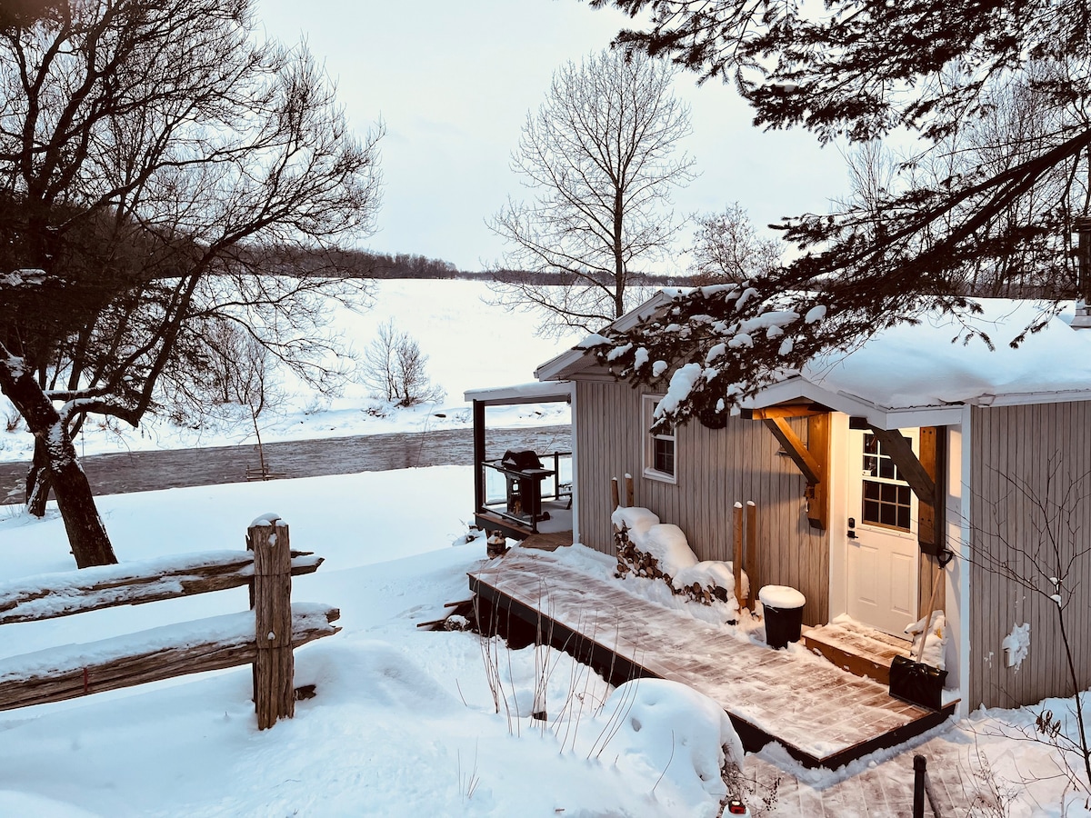 A rustic chalet is seen from an elevated angle, with snow blanketing the ground. A wooden deck leads to the entrance, while a gas grill is positioned nearby. Leafless trees frame the view of the serene winter landscape and the river beyond.