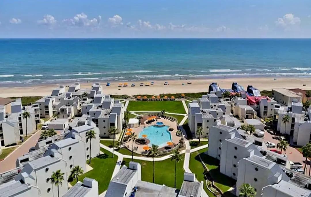 An aerial view of a resort showcases white buildings surrounding a central pool area. Lush green landscaping leads to the beachfront, where colorful beach chairs and umbrellas are positioned along the shore. The blue ocean extends into the distance under a clear sky.