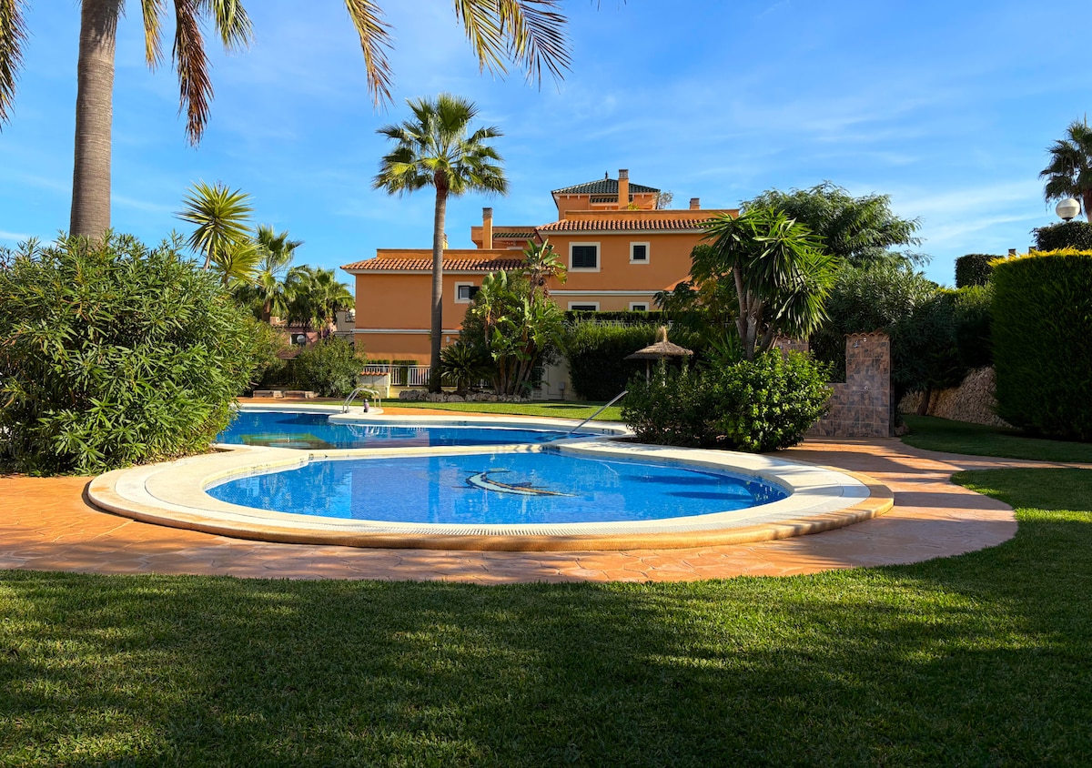 A large communal pool is surrounded by lush greenery and palm trees. The pool area features a separate jacuzzi section, and sunbeds can be seen around the pool. The buildings in the background provide a sense of privacy, complementing the tranquil landscape.