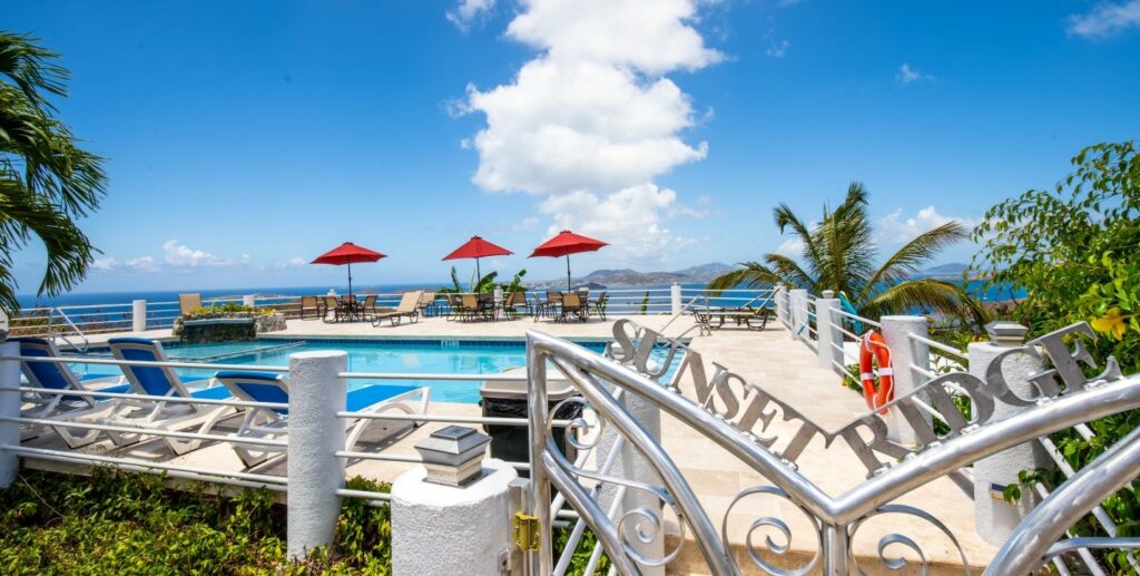 A view of the villa's pool area reveals several lounge chairs with red umbrellas overlooking the ocean. The blue sky is dotted with clouds, while lush greenery frames the setting, enhancing the atmosphere of relaxation and leisure.
