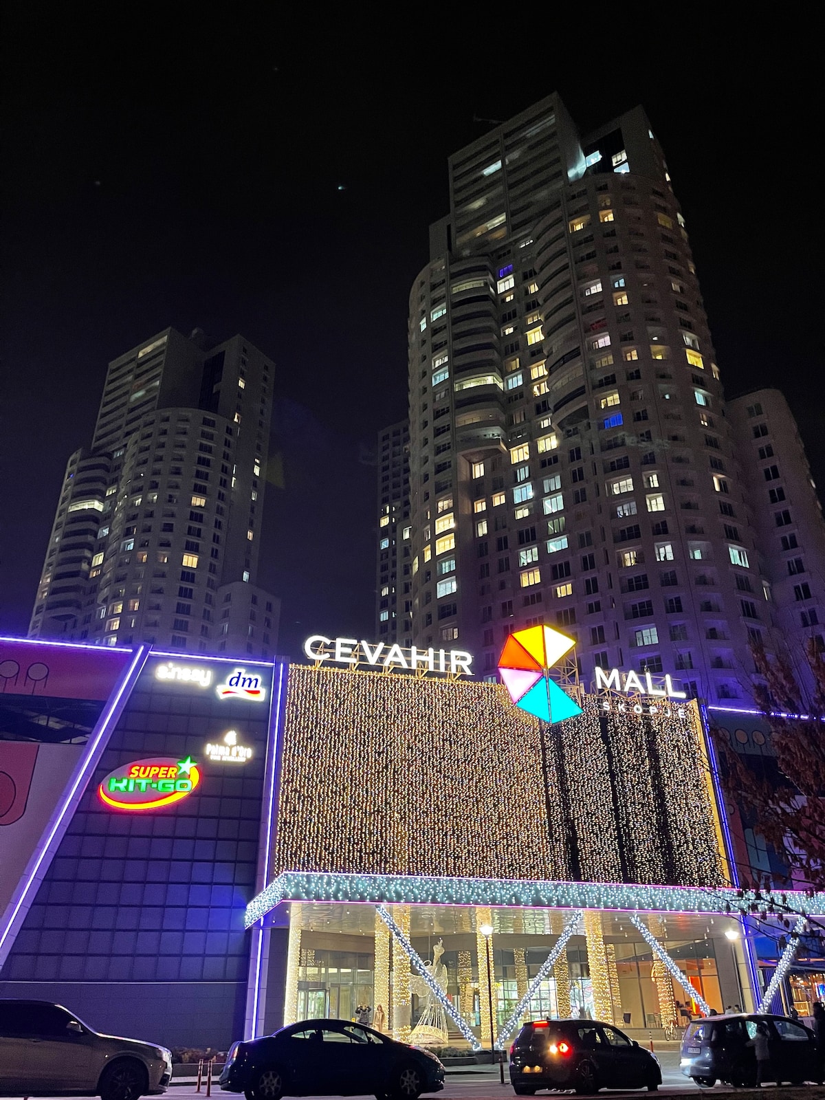 The Cevahir Mall exterior is illuminated at night, showcasing a vibrant façade adorned with colorful lights. The mall's sign is prominently displayed, while tall residential towers rise in the background. A few parked cars can be seen in front of the entrance.