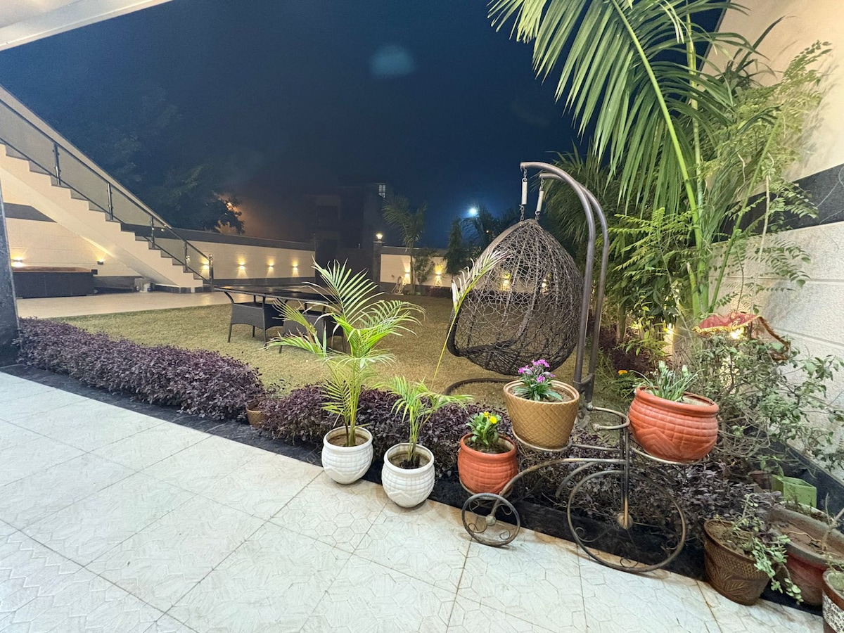 A landscaped garden area is featured, showcasing soft lighting against lush greenery. Potted plants and flowers are arranged along the edges, while a decorative hanging chair is positioned on a patio with outdoor seating visible in the background. A staircase leads to an upper level.