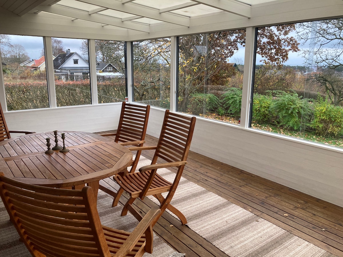 A sunlit porch is surrounded by large windows, showcasing a scenic view of the outdoors. Wooden furniture, including a round table and several slatted chairs, occupies the space, complemented by a textured area rug on the wooden floor.