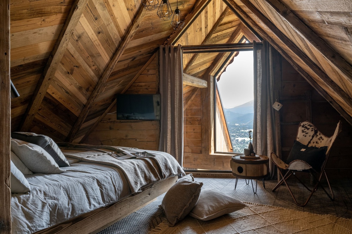The bedroom features a spacious bed with neutral bedding, positioned under a sloped wooden ceiling. A large window offers a view of the surrounding mountains. A cozy armchair is placed beside a small table, and a soft cushion rests on the floor.