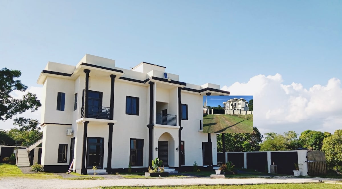 A modern two-story building is showcased, featuring a clean, white exterior with contrasting black columns. Large windows allow for ample natural light, while a landscaped garden with green foliage is visible in the foreground, enhancing the serene ambiance of the property.