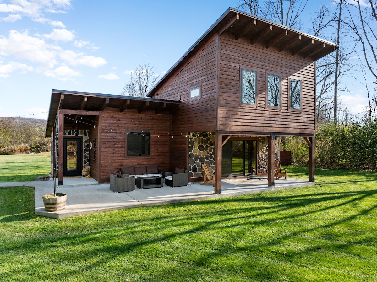 A modern wooden structure with large windows is situated on a grassy area. A covered patio is adorned with comfortable seating, inviting outdoor relaxation. The scenic backdrop features trees and open space, enhancing the peaceful ambiance of the retreat.