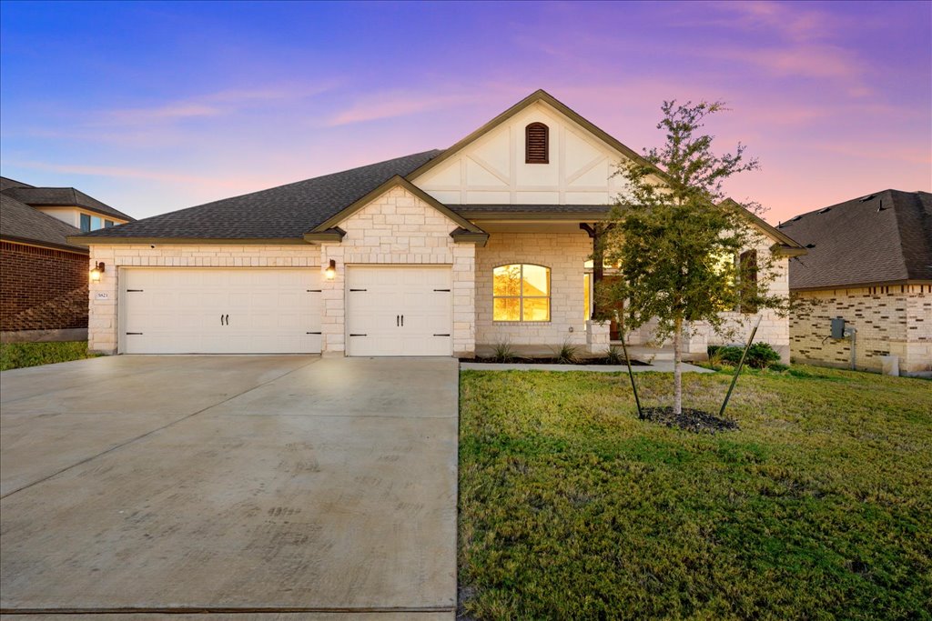 The exterior of a modern, single-story house is shown, featuring a welcoming facade of light-colored stone and white accents. A driveway leads to a two-car garage, while well-maintained grass and a small tree enhance the front yard, set against a vibrant sunset sky.