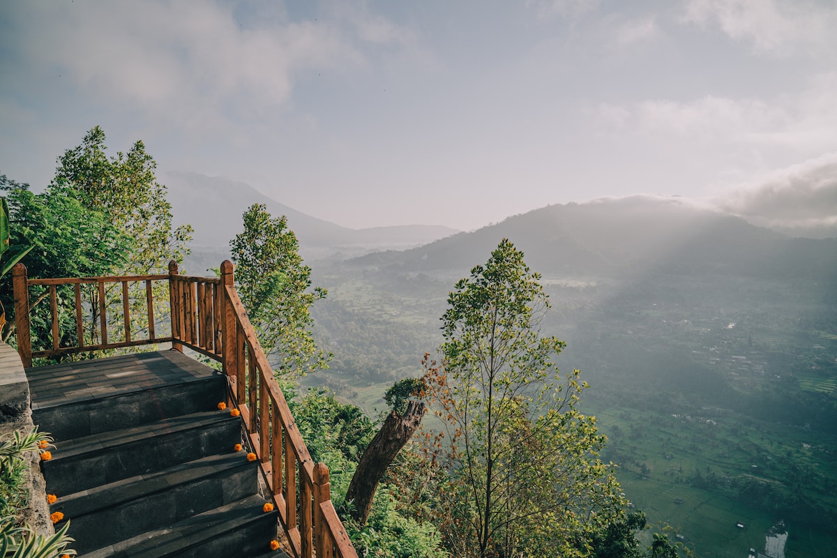 A wooden staircase ascends towards an expansive view of rolling hills and lush valleys. Sunlight filters through a light mist, creating a serene atmosphere. The surrounding trees provide a natural frame for the scenic landscape, with hints of vibrant orange flowers along the staircase.