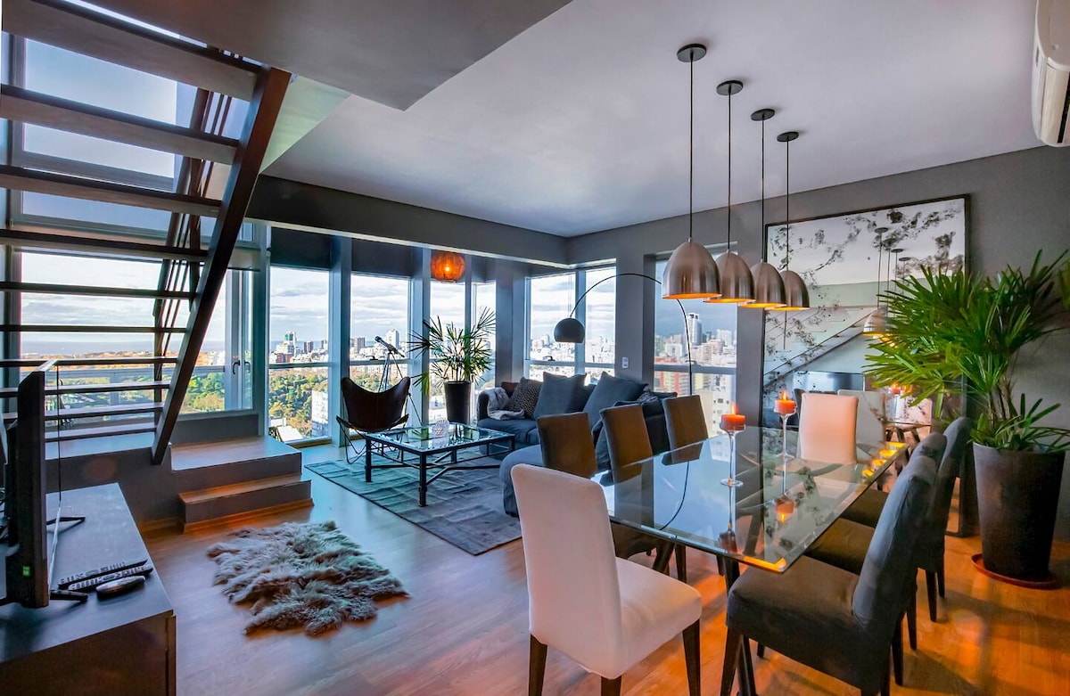 A spacious living area is illuminated by natural light, featuring large windows with panoramic city views. A modern dining table accommodates several chairs, while a comfortable seating arrangement includes a sofa and decorative elements like plants. The staircase adds an architectural dimension to the space.