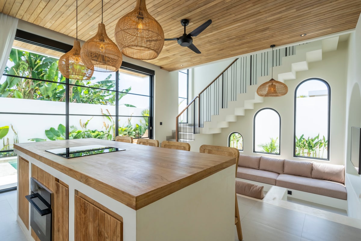 A spacious open-plan living area features a large central kitchen island made of wood. Natural light fills the space through expansive glass doors, displaying lush greenery outside. Unique pendant lights hang from the ceiling, complementing the wooden accents and modern design throughout the villa.