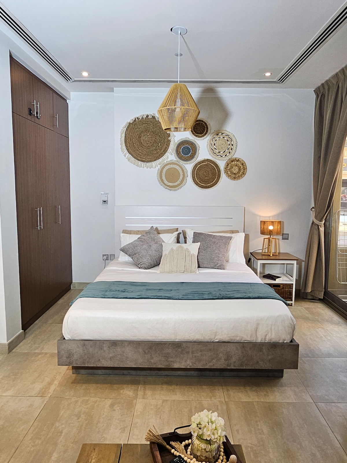 A modern bedroom features a plush queen bed adorned with muted textiles. Wall decorations of woven patterns add texture, complemented by a bedside lamp. A built-in wardrobe offers storage solutions, and natural light enters through large windows, enhancing the space’s openness.