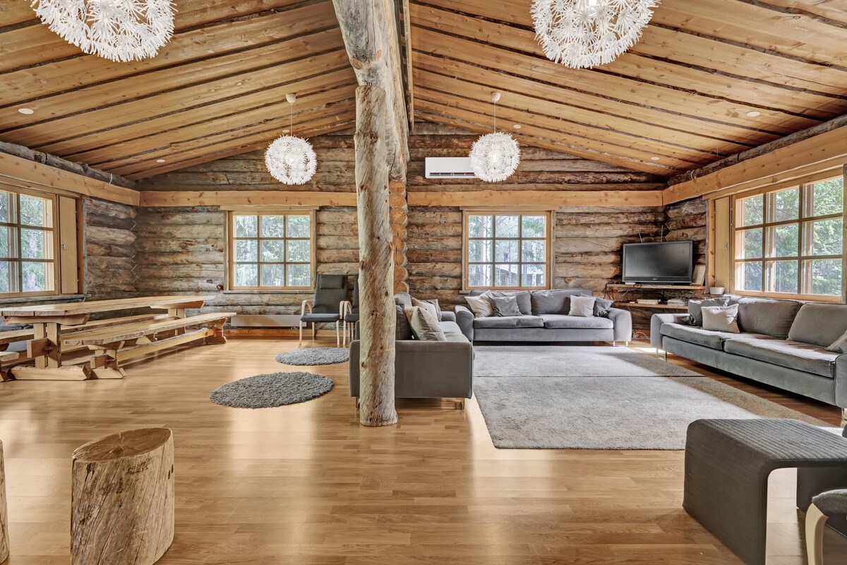 A spacious living area features high wooden ceilings with exposed beams. Comfortable seating includes multiple gray sofas arranged around a large area rug. A long wooden dining table is positioned to one side, accompanied by natural light filtering through generous windows.