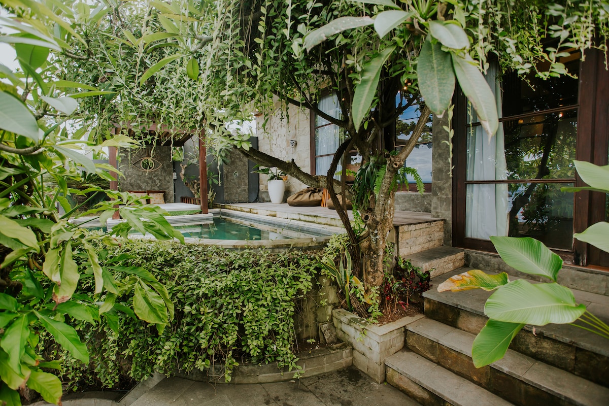 A private pool area is framed by lush greenery and tropical plants, offering a serene environment. Stone steps lead to the pool, which is partially visible. The surrounding foliage creates a secluded atmosphere, inviting relaxation while blending with the natural landscape.