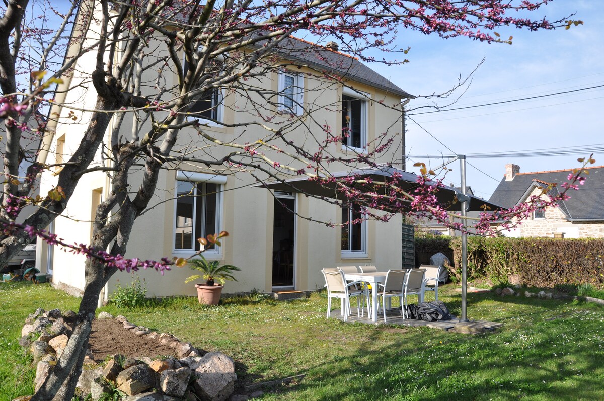 A two-story house is portrayed in a garden setting. The exterior features a pale yellow facade and large windows. A dining table with six chairs is visible on the terrace, surrounded by greenery and flowering trees. A stone border outlines a grassy area.