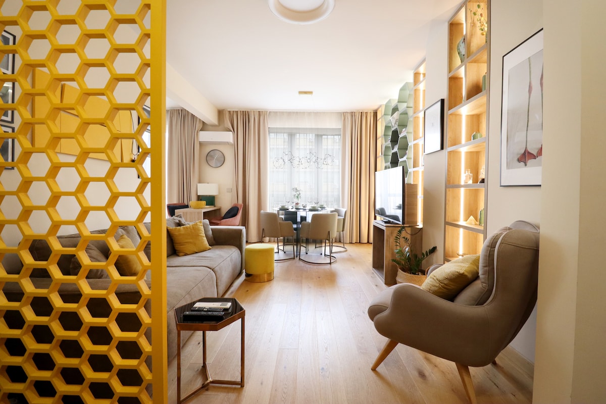 A stylish living area is depicted, featuring a cozy sofa and an accent chair. Natural light flows through the window, highlighting decorative shelving and soft furnishings. A dining table for four is visible in the background, surrounded by warmly lit shelves and subtle plants.
