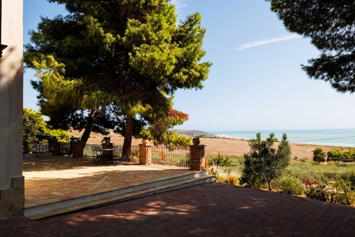 An outdoor terrace is shown with a paved surface, surrounded by lush green trees. A view of the sea and rolling hills is visible in the background, providing a serene atmosphere for relaxation.