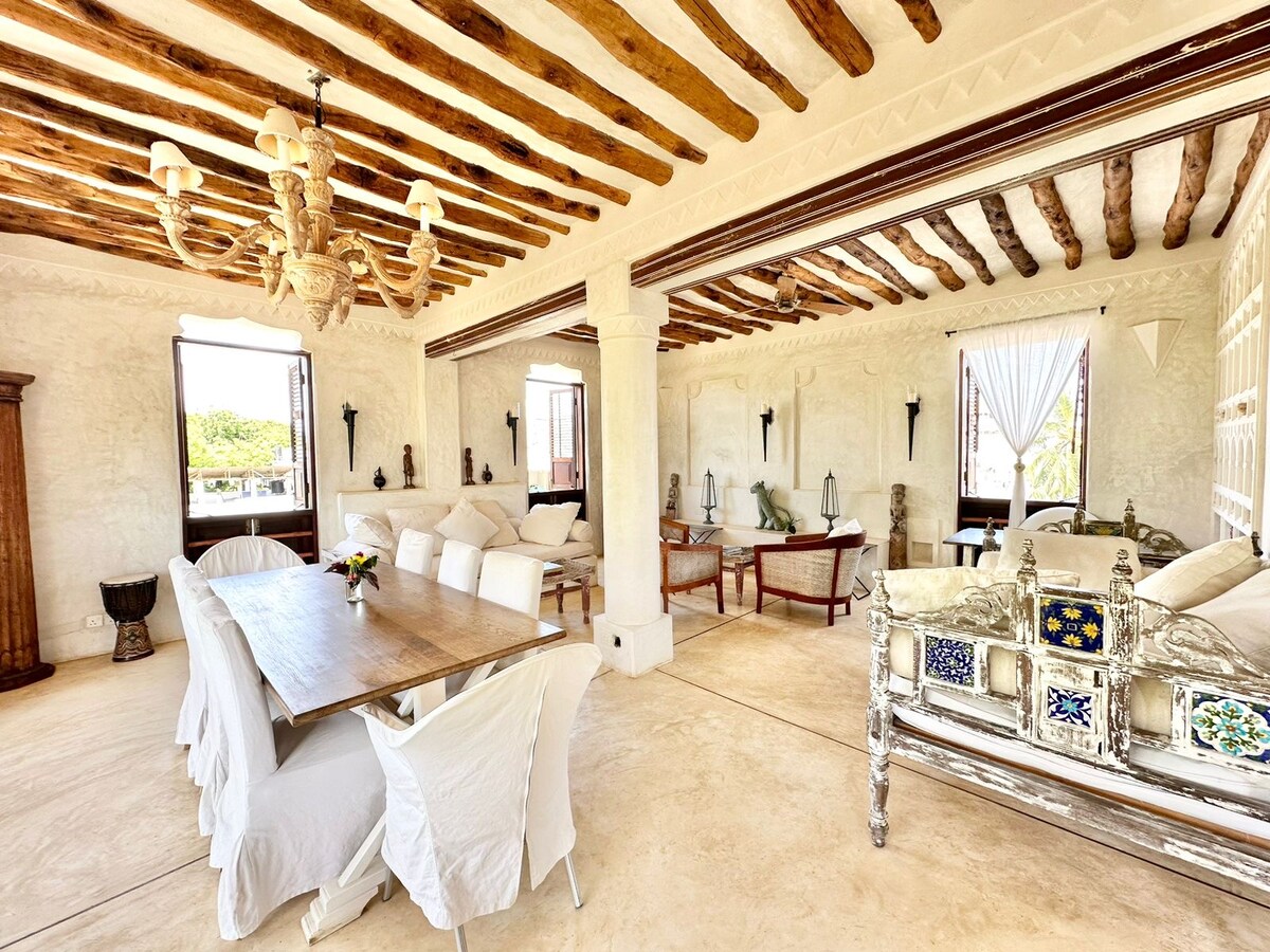 An airy living and dining area features high, exposed beam ceilings and elegant furnishings. A large dining table is surrounded by white chairs, while seating areas with plush cushions invite relaxation. Light filters through windows, highlighting the subtle textures of the walls.