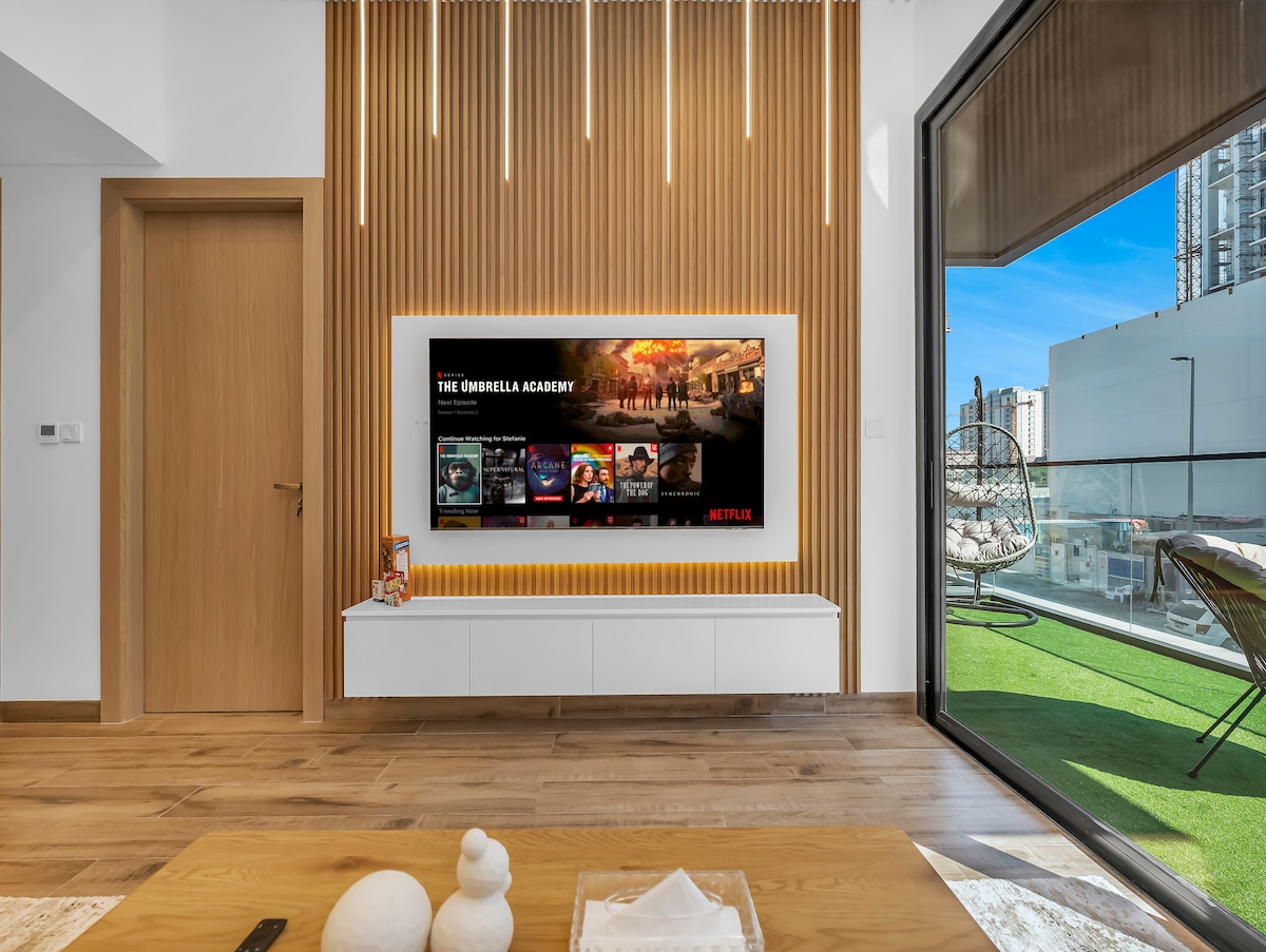 A modern living area features a sleek media wall with a large Samsung Smart TV displaying various shows. Soft wooden accents and natural light filter through large windows, while an outdoor balcony with green turf is visible in the background.