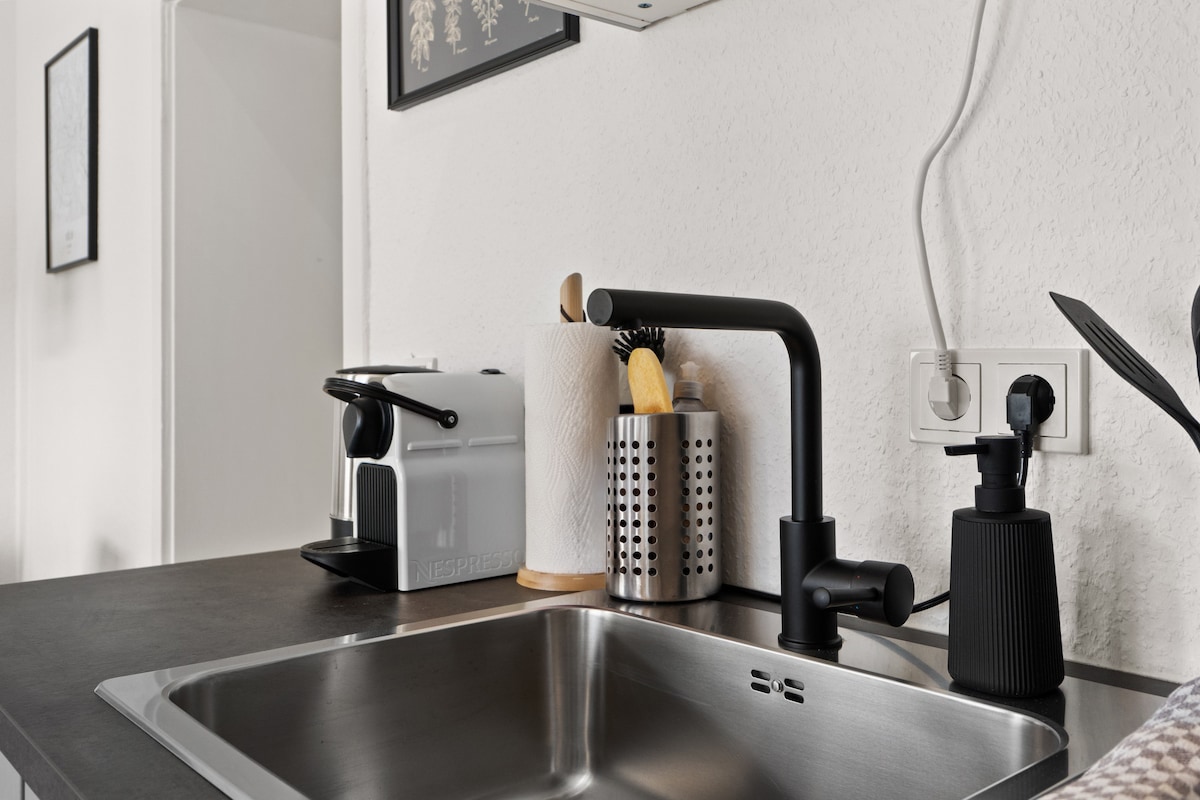 The modern kitchenette is equipped with a stainless steel sink, a sleek black faucet, and organized kitchen utensils in stainless steel holders. A coffee machine is visible alongside essential cooking tools, creating a functional and streamlined cooking space.